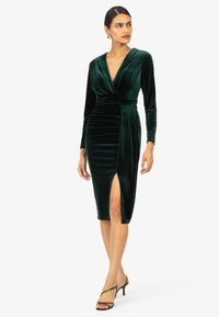 Groene fluwelen midi-jurk met lange mouwen, diepe V-hals en een zijsplitting. Heeft geplooide stof bij de taille voor een getailleerd ontwerp.