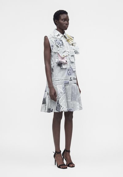 Γυναίκα που φοράει αμάνικο φόρεμα μέχρι το γόνατο με τύπωμα εφημερίδας, μεγάλο πολυεπίπεδο γιακά και μαύρα πέδιλα με λουράκι στον αστράγαλο και ψηλό τακούνι.