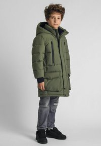 Abrigo de invierno acolchado verde con capucha, con bolsillos negros con cremallera, junto con jeans gris desgastados y zapatillas negras.