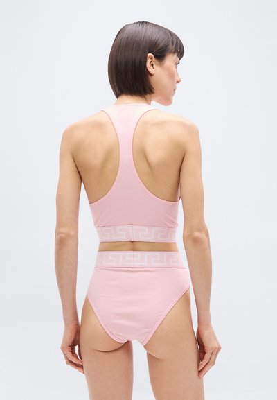 Γυναίκα με κοντά καστανά μαλλιά, φορώντας ροζ ψηλόμεσο εσώρουχο και τοπ χωρίς μανίκια με γεωμετρική ζώνη, με την πλάτη γυρισμένη σε λευκό απλό φόντο.