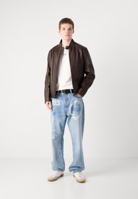 Brown leather jacket over a white t-shirt, paired with light blue jeans featuring graphic designs. White shoes with tan soles complete the look.