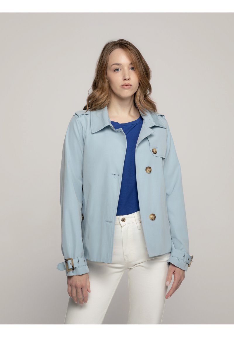Lichtblauwe jas van een glad materiaal, met een kraag, gouden knopen en gespdetails op de mouwen. Draagt over een blauwe top en witte broek.