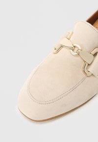 Beige suede loafer with round toe, decorative gold-tone hardware, stitched detailing, and a smooth texture on the upper.