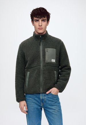 Jeune homme aux cheveux bouclés portant une veste polaire verte zippée avec poches avant et un jean bleu, debout sur un fond blanc.