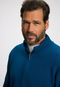Geverfde, blauwe ritssweater met een geribbeld kraagje, met een halflange rits en gelaagde look over een witte onderhemd.