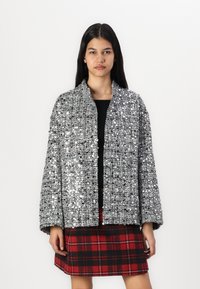 Veste argentée à sequins avec une coupe ample et des manches larges, présentant une texture côtelée. Portée par-dessus un haut noir et une jupe à carreaux.