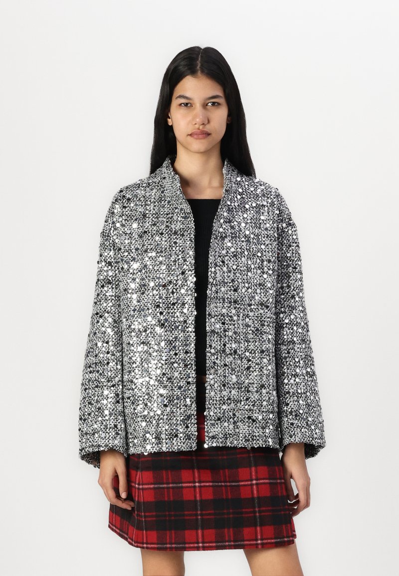 Veste argentée à sequins avec une coupe ample et des manches larges, présentant une texture côtelée. Portée par-dessus un haut noir et une jupe à carreaux.