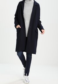 Langer marineblauer Mantel mit lockerer Passform, zwei Fronttaschen, Knopfverschluss und Revers, kombiniert mit einem hellgrauen Pullover und Jeans.