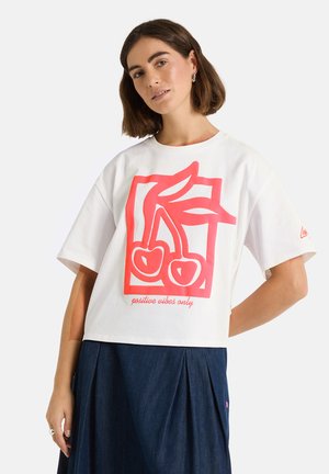 Woman with short brown hair wearing a white T-shirt with red cherry graphic and "positive vibes only" text, paired with a dark blue skirt.