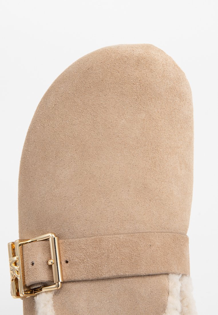 Chausson en suede beige avec un bout arrondi, présentant une boucle en métal doré et un doux revêtement en fourrure le long de l'ouverture. Texture lisse dans l'ensemble.