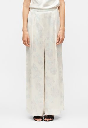 Kobieta w jasnych, kwiatowych spodniach z wysokim stanem i szerokimi nogawkami oraz w przezroczystych czarnych szpilkach z odkrytymi palcami, stojąca na jednolitym tle.