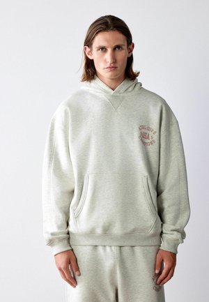 Young man with shoulder-length hair wearing a light gray hoodie and matching sweatpants with red text on the chest and thigh.