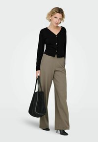 Ženska, oblečena v črno V-izrezno zgornjo s sponkami, taupe široke hlače, črne škornje s peto, drži veliko črno torbo na nevtralni podlagi.