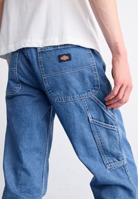 Pantalones cargo de mezclilla azul claro con múltiples bolsillos, un acabado texturizado y una etiqueta en el bolsillo trasero. Ajuste de pierna ancha con diseño casual.