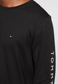 Schwarzes Langarmshirt aus Baumwolle. Mit einem kleinen gestickten Logo in Rot, Weiß und Blau sowie dem Schriftzug "TOMMY" in Weiß auf dem Ärmel.
