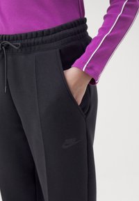 Person som bär svarta Nike sweatpants med handen i fickan och en lila långärmad tröja med vit rand på ärmen.