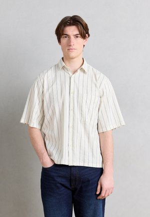 Ung mann med brunt hår iført en kortermet beige skjorte med tynne, vertikale striper og mørkeblå jeans, stående med den ene hånden i lommen.