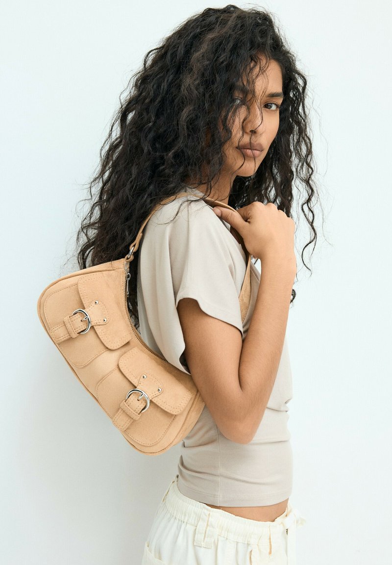 Femme aux longs cheveux bouclés, portant un haut beige et un pantalon blanc, tenant un sac à main beige avec des boucles sur son épaule.