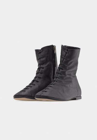 Μαύρα μαλακά δερμάτινα μποτάκια αστραγάλου με κορδόνια μπροστά, πλαϊνό φερμουάρ και επίπεδες ανοιχτό καφέ σόλες, εμφανιζόμενα σε απλό λευκό φόντο.