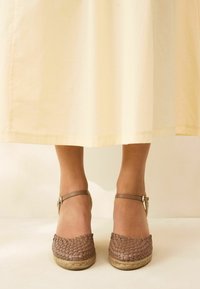 Woven brown espadrilles with a beige jute sole and adjustable ankle straps, positioned beneath a cream-colored skirt.
