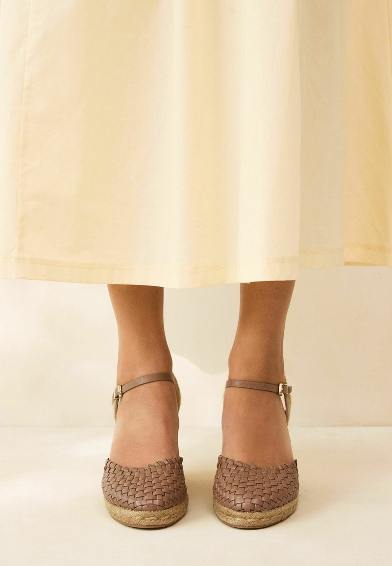 Woven brown espadrilles with a beige jute sole and adjustable ankle straps, positioned beneath a cream-colored skirt.