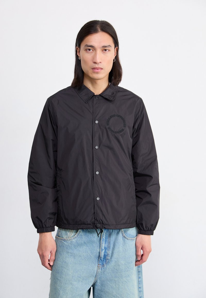 Black rain jacket with a collar, snap buttons, elastic cuffs, and a logo in matching black on the left side. Worn with light blue jeans.