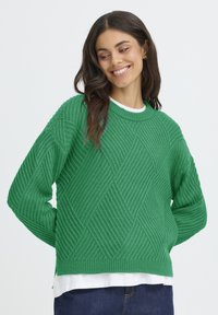 Pulover verde tricotat, cu un model în diamant, textură ribbed și mâneci lungi. Purtați deasupra unei cămăși albe, cu o croială lejeră.