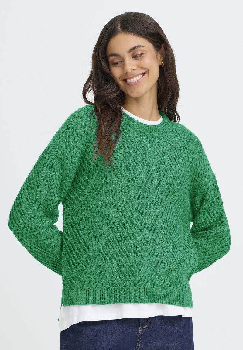 Pulover verde tricotat, cu un model în diamant, textură ribbed și mâneci lungi. Purtați deasupra unei cămăși albe, cu o croială lejeră.