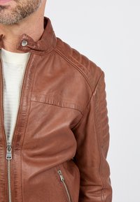 Veste en cuir marron avec un col montant, fermeture éclair et accents matelassés aux épaules. Elle est dotée de poches latérales zippées et d'une texture lisse.