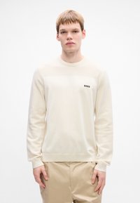 Giovane uomo che indossa un maglione lavorato a maglia color crema con dettagli a coste e il logo "BOSS", abbinato a pantaloni beige, in piedi su uno sfondo bianco.