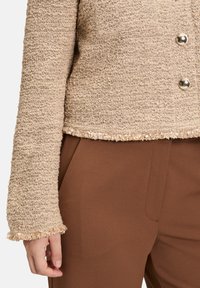 Giacca beige in tessuto con design accorciato, caratterizzata da un orlo sfrangiato e bottoni in argento. Abbinate a pantaloni marroni.
