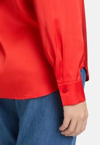 Rotes, seidiges Langarmhemd mit Knopfmanschette, das eine glatte Textur und eine lockere Passform aufweist, kombiniert mit blauen Denim-Hosen.