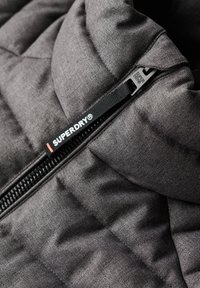 Grijze gewatteerde jas met een hoge kraag en een rits aan de voorkant. De rits heeft een zwart merklabel met de tekst "SUPERDRY". Textuurstof.