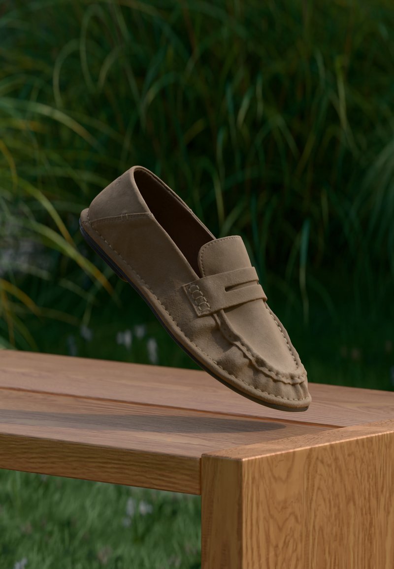 Καφέ μοκασίνι από σουέντ που αιωρείται πάνω από ένα ξύλινο παγκάκι με πράσινα φύλλα θολωμένα στο φόντο.