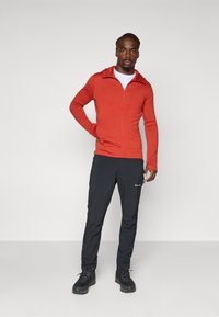 Rode hoodie met rits, witte shirt eronder, zwarte broek met zijzak, zwarte schoenen. Glad materiaal, getailleerd ontwerp, lange mouwen, casual uitstraling.