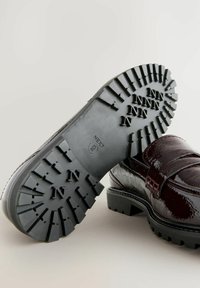 Chaussure en cuir verni bordeaux avec une semelle en caoutchouc noir épaisse dotée de profondes empreintes et portant la mention "NEXT" en relief. Accentuée d'un large détail de bride.