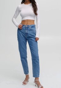 Crop top blanc à manches longues associé à un jean taille haute bleu clair et des sandales à talons hauts transparentes à brides. Design simple, silhouette ajustée.