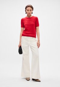 Piros, rövid ujjú pulóver, texturált felülettel, fehér, bő szabású nadrággal párosítva. Fekete clutch táska és bézs, blokk sarkú cipő.