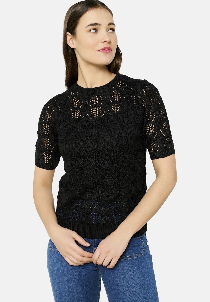 Melna adīta īsām piedurknēm džemperis ar caurspīdīgu rakstu, apaļa kakla izgriezums un teksturēti elementi. Apvienots ar ziliem džinsiem.