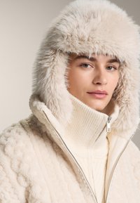 Osoba oblečená v krémovej bunde s kapucňou lemovanou kožušinou a hrubom rebrovanom roláku, smerujúca dopredu s neutrálnym výrazom tváre.