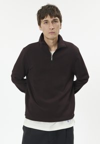 Burgunderfarbener Zip-Hoodie mit hohem Kragen, aus weichem Stoff, mit enganliegender Passform und locker sitzenden Ärmeln, über einem weißen Shirt getragen.