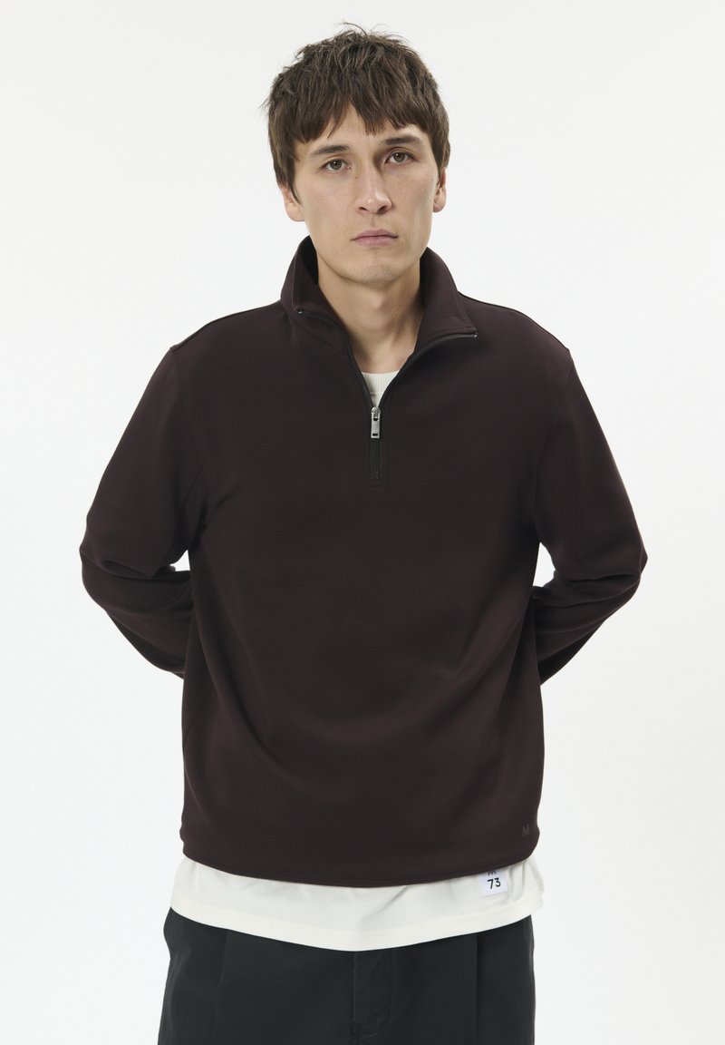 Burgunderfarbener Zip-Hoodie mit hohem Kragen, aus weichem Stoff, mit enganliegender Passform und locker sitzenden Ärmeln, über einem weißen Shirt getragen.