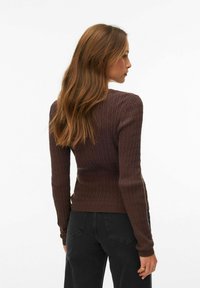 Femme aux longs cheveux bruns portant un pull en maille torsadée marron et un jean noir, debout et regardant vers la droite sur un fond blanc.