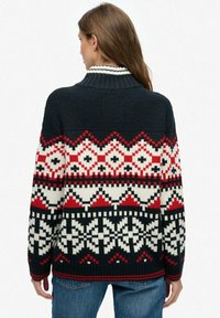 Vrouw in een gebreid sweatshirt met rode, witte en zwarte geometrische patronen, staande met haar rug naar de camera gekeerd.