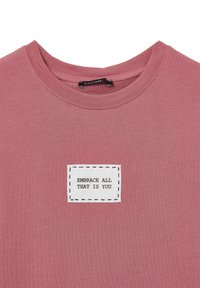 T-shirt in cotone rosa con scollo rotondo e una patch rettangolare bianca con il testo "ABBRACCIA TUTTO CIÒ CHE SEI" in nero.