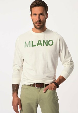 Langärmliges Cremeshirt mit grünem "MILANO"-Grafik. Kombiniert mit olivgrüner Hose. Brauner Gürtel und Armband am Handgelenk sichtbar.
