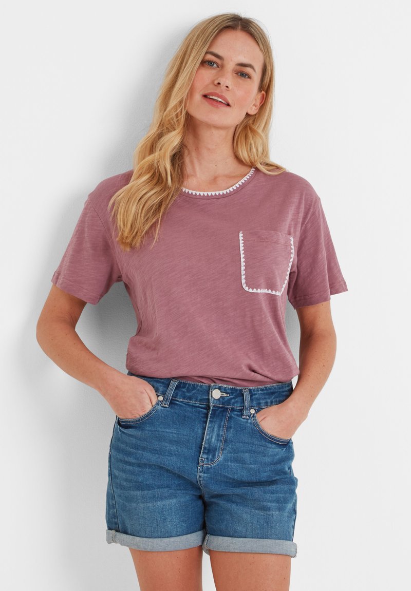 Woman with long blonde hair wearing a mauve t-shirt with white trim pocket and rolled-up denim shorts, standing with hands in pockets.