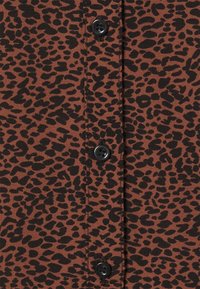 Tissu marron avec un motif de taches noires irrégulières, présentant des boutons noirs le long du centre. Texture lisse avec un léger éclat.