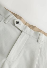 Pantalon en tissu gris clair texturé avec un bouton noir et une ceinture intérieure beige, avec passants de ceinture et détail de couture avant.