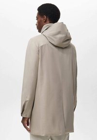 Mand iført en beige hættejakke, der vender væk mod en ensfarvet lys baggrund, viser ryg- og sideprofil.